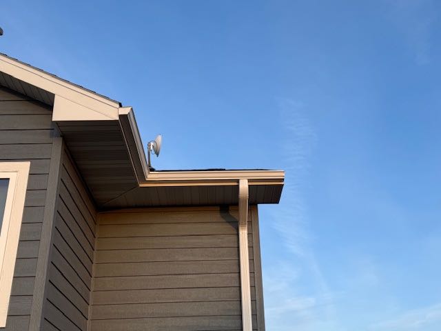 The image shows the corner of a gray house with clapboard siding and a satellite dish on the roof, set against a clear blue sky. The scene feels calm and serene.