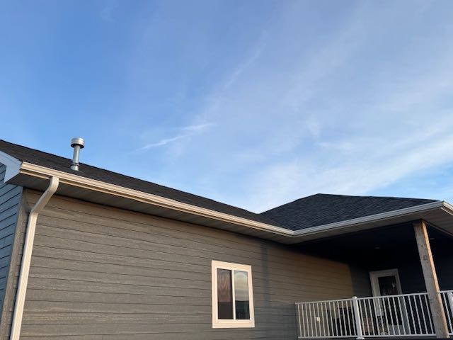 Single-story house with gray siding and a dark roof against a clear blue sky. There is a small chimney, a white-framed window, and a railing.