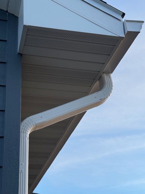 Close-up of a house's roof edge showing a white gutter system against blue siding and a light sky. The scene conveys a clean, orderly appearance.
