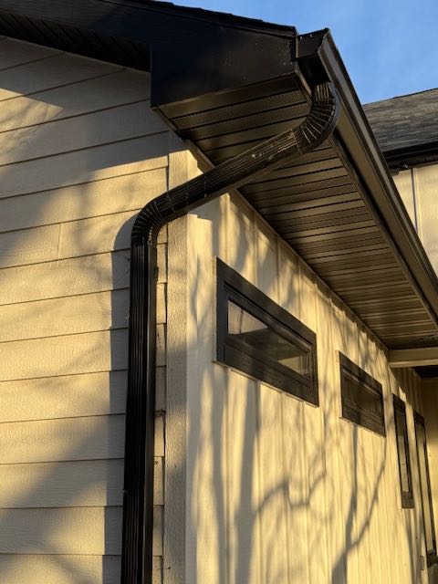 The image shows a beige house exterior in evening light, featuring black gutters, soffit, and narrow horizontal windows. Tree shadows create a calm atmosphere.