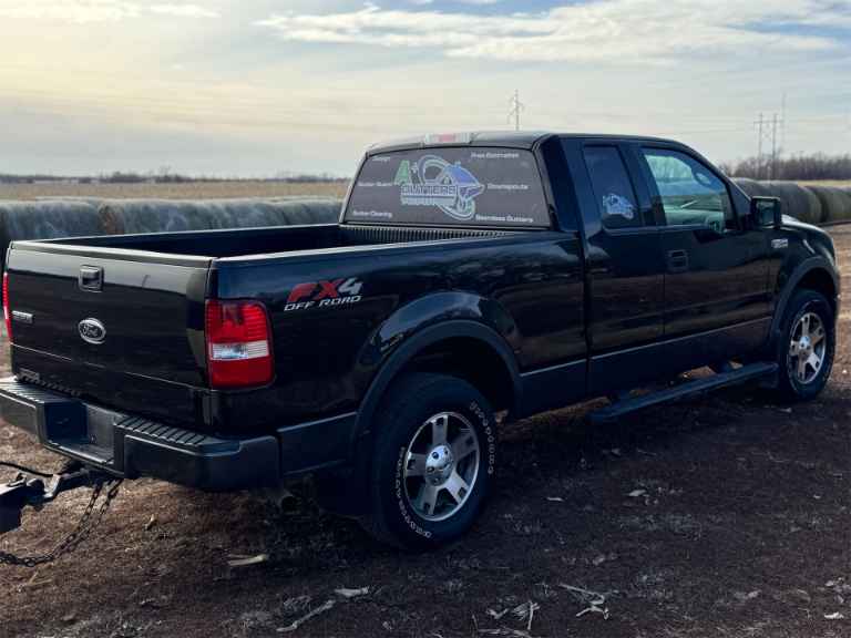 A black Ford FX4 Off Road truck is parked on dirt near large hay bales. The sky is cloudy, and the truck's rear window features a branded decal.